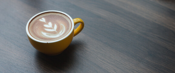 Close-up of hot cappuccino with beautiful latte art in yellow ceramic cup on wooden table.
