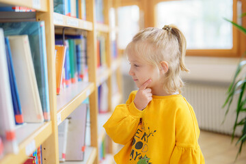 Young girl thoughtfully exploring a library filled with colorful books while wearing a bright yellow sweater