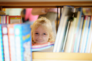 Child peeking through a bookshelf in a library during daytime activities with other visitors in the background