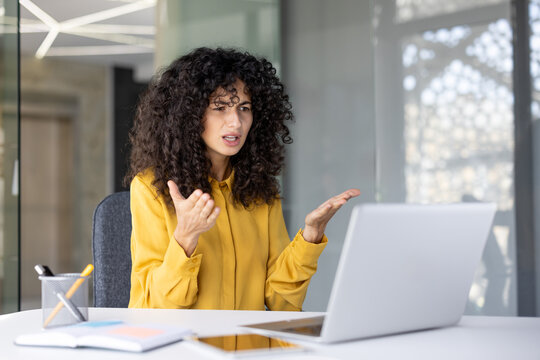 A woman with curly hair appears frustrated during a video call, gesturing with her hands while seated in front of a laptop.