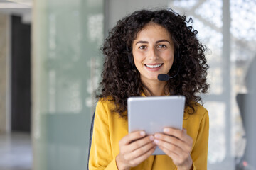 A smiling woman wearing a headset holds a tablet, appearing ready for customer service in a modern...