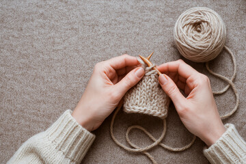 A person hands is knitting a cozy beige swatch with wooden needles, surrounded by a ball of yarn on a textured surface.