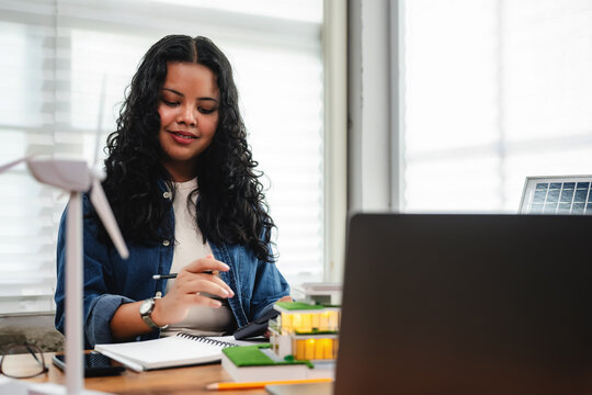 Female architect designing sustainable house with wind energy model, Eco-friendly building model with wind turbine in modern office, Architecture student creating smart green home design