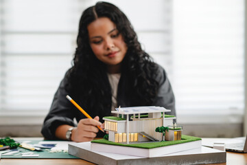 Smiling female architect or designer working with modern house model placed on table in bright and...