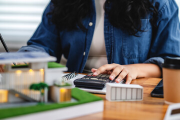 Fototapeta premium Woman calculates with a calculator beside a modern eco-house model, notebook, and coffee cup, representing sustainable planning, budgeting, and green architecture projects.
