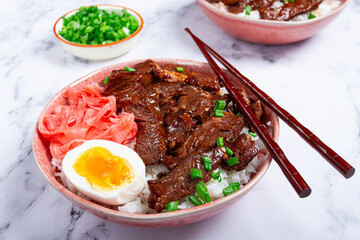 Gyudon Japanese beef with rice, pickled ginger, boiled egg slice, garnished with green onions, in a bowl with chopsticks,