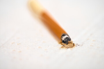 Close-up of cigarette resting against white fabric, leaving burn mark. Soft lighting highlights damage, contrasting with brown and white tones. Shallow depth of field focuses on burn area