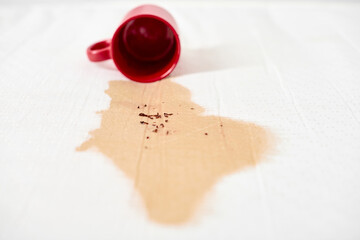 Red mug lies tipped over, spilling coffee onto white textured surface. Brown liquid creates...
