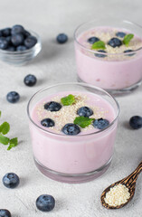 Thick yogurt with blueberries and sesame seeds in glasses, as well as in wooden spoon on gray background