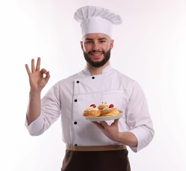Happy confectioner in uniform holding delicious profiteroles with strawberries and showing ok gesture on white background