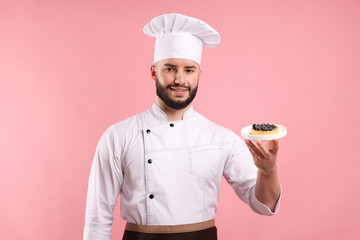 Happy confectioner in uniform holding tart with blueberries on pink background