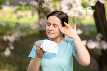 Woman sneezes into handkerchief in blooming garden wiping away tears, symptoms of seasonal...