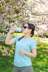 Fototapeta premium Young woman wearing sunglasses drinking water from bottle outdoors in blooming spring park