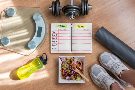 Fitness enthusiast preparing for workout with a healthy salad, dumbbells, yoga mat, scale, and water bottle, following a personalized meal plan for a balanced lifestyle