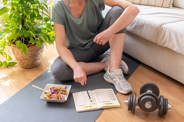 Mature woman sitting on a yoga mat at home, pointing at her weekly meal and workout plan while...