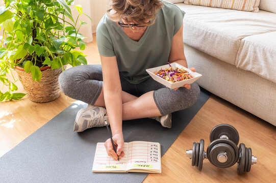 Sitting on a yoga mat, a mature woman writes down her meal plan while holding a healthy salad, with dumbbells nearby, symbolizing her commitment to a balanced lifestyle