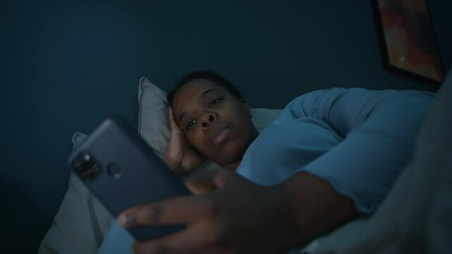 Young Black woman lying in bed in dark room, holding smartphone and scrolling through social media while suffering insomnia during night
