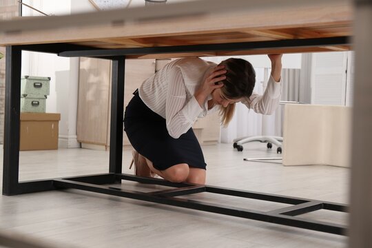 Scared young woman hiding under office desk during earthquake