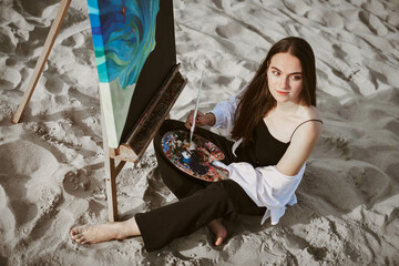 Young female artist sits barefoot on sandy beach, painting on canvas. Serene environment inspires...