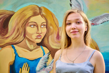Young blonde female standing confidently before colorful mural. Artwork depicts woman, bird, blue dress. Sunny day, soft shadows enhance details beautifully