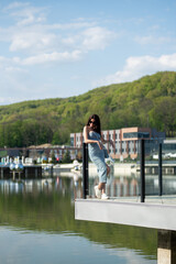 Fototapeta premium Young stylish woman in a denim dress sitting on a concrete bench outdoors on a sunny day. She is wearing sunglasses and white sneakers, enjoying the warm weather in an urban park setting.