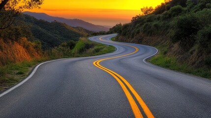 Fototapeta premium A winding asphalt road under a golden sunset stretches beside a slope with vegetation and an open sea horizon, highlighted by yellow and white road markings for scenic travel imagery.