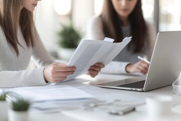 Two women collaborate on documents, reviewing papers and using a laptop for efficient teamwork.
