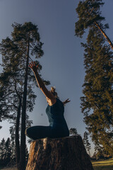 A woman practicing yoga performs a sitting in lotus position on a tree stump, meditating alone in...