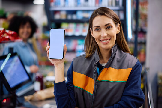 Happy supermarket worker showing store app on mobile phone at cash register.