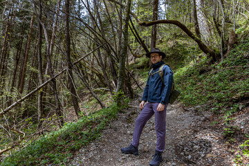 A young man of about thirty in a wide-brimmed hiking hat and clothes, with a backpack, stands on a picturesque Alpine path, in the spring in Austria © Vlad