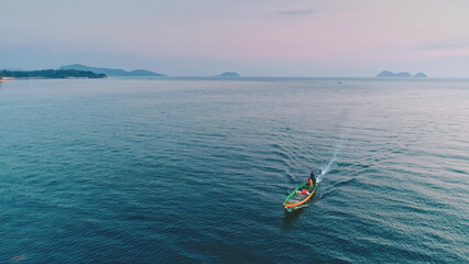 Aerial view of a small fishing boat navigating the calm waters of a tropical sea at sunset, with islands and coastline visible in the distance, creating a serene and picturesque scene