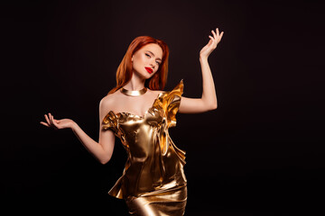 Elegant woman in a golden dress with red hair and glamorous makeup posing stylishly against a dark...
