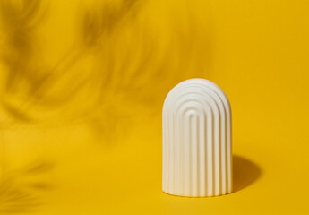 A small, white ceramic decorative arch with vertical ribbed texture stands on a bright yellow surface with blurred leaf shadows.