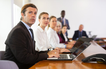 Fototapeta premium Serious middle-aged white brunette businessman sitting among colleagues at conference table in meeting room, intently listening to presentation and taking some notes on laptop during corporate