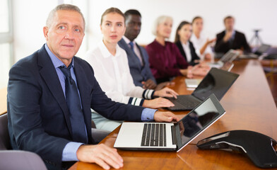 Fototapeta premium Positive middle aged businessman in suit sitting at table with his colleagues, attending meeting and using laptop.