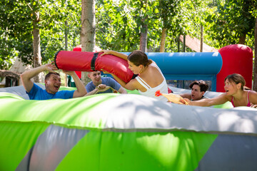 Adults having fun on inflatable amusement playground. Emotional young girl fighting off her friends with inflatable log while they trying to filch toy chickens