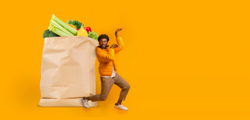 A man joyfully poses next to a large paper bag filled with fresh vegetables and fruits, celebrating the grocery shopping experience during the new normal. Bright yellow background adds vibrancy.