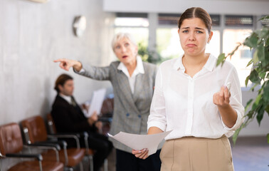 Upset young woman in business clothes with documents in her hands being criticized in office
