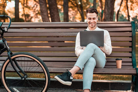 Work outdoors. Freelance businessman typing at laptop and sitting on bench with takeaway coffee with bike in park, panorama, copy space