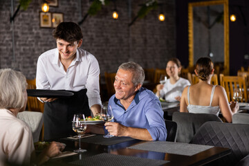 Elderly male and female colleagues have fun chatting and spending time in restaurant after working day. Waiter guy serves wine order and wishes cafe visitors pleasant appetite and good time