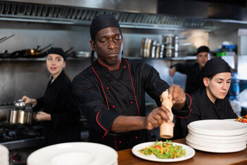 Focused young adult man chef seasoning salad before giving it out on order station in restaurant kitchen