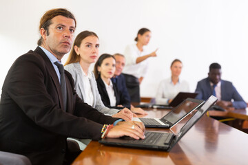 Focused mature white business man in formal suit sitting with colleagues in conference room and absorbedly listening to speaker's presentation during corporate group meeting