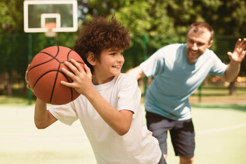 Active Leisure Concept. Portrait of cheerful father and son playing basketball together, man trying to catch the ball, boy running from him, selective focus, blurred background