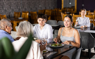 In cozy restaurant, young family and mature parents enjoy festive lunch, celebrating important event. Company chat and taste wine, enjoying pleasant atmosphere and good company.