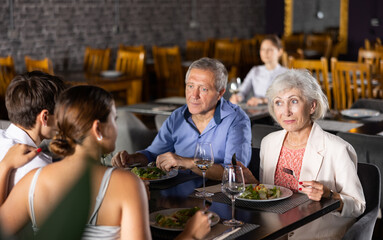 Senior parents enjoying delicious food and pleasant conversation with adult children over dinner in restaurant. Happy carefree people gathered around table for family celebration..