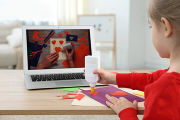 Little girl making handmade project with color paper and glue by video tutorial at table indoors