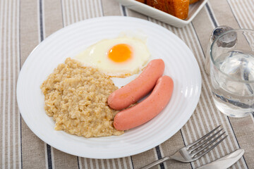 Two sausages with a fried egg and oatmeal on a white plate. Breakfast option with eggs and porridge next to cutlery and a glass of water