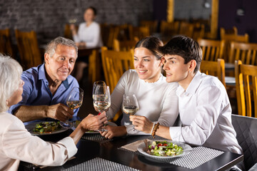 In cozy restaurant, young family and mature parents enjoy festive lunch, celebrating important event. Company chat and taste wine, enjoying pleasant atmosphere and good company.