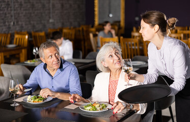 Adult woman waiter serving wine to couple of elderly man and woman in restaurant