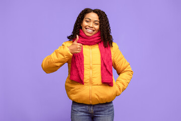 Black Lady Gesturing Thumbs-Up Smiling To Camera, Approving And Recommending Something Standing Over Blue Studio Background, Wearing Winter Clothes. Like, Thumbs Up Gesture Concept.